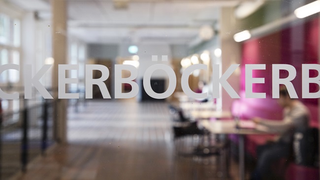 The Swedish word for books, "böcker", is written on a glass wall. A pink sofa in a corridor shows on the other side of the wall. Photo.